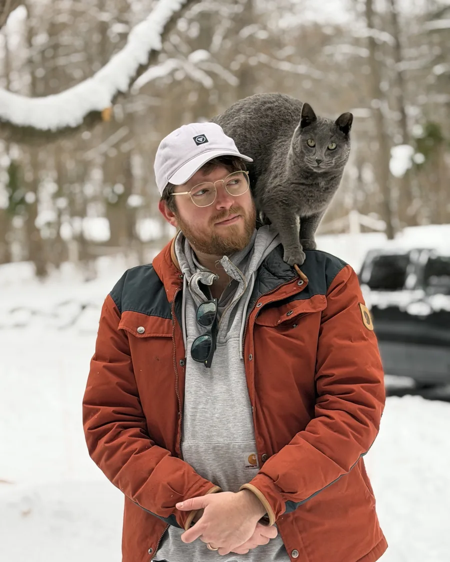 Erik outside in the snow in a rust-colored jacket and white cap, with a gray barn cat standing on his shoulder.